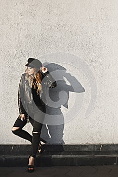 Modish blonde model girl in a leather jacket, ripped jeans, and black cap standing outdoors against the urban wall in photo