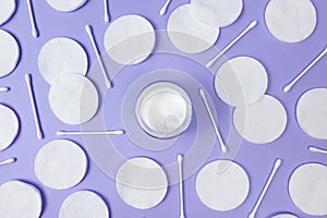 Moisturizer cream in open glass jar, cotton pads and cotton sticks on textured light purple background. Top view. Natural bio