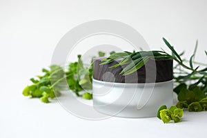 Moisturizer facial or eye cream in frosted glass jar with wooden cup and meadow herbs and leaves on beige background. Concept eco