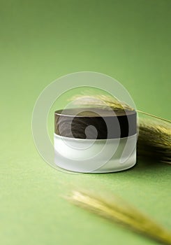 Moisturizer facial or eye cream in white frosted glass jar with wooden cup mockup and spikelet on yellow-green background
