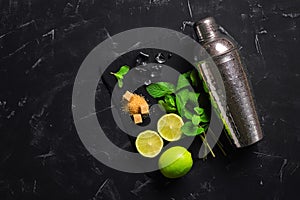 Mojito cocktail ingredients. Mint leaves, lime, ice cubes, brown sugar, and a shaker. Black stone background. Top view, flat lay,