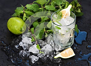 Mojito cooking set. Bunch of fresh mint, lime, chipped ice and coctail glass over black slate stone backdrop