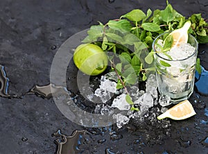 Mojito cooking set. Bunch of fresh mint, lime, chipped ice and coctail glass over black slate stone backdrop, copy space