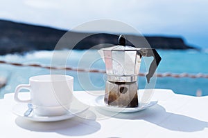 Moka pot espresso maker and cup of coffee on table with coastline and ocean in background