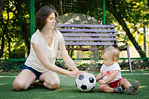 Mom and baby roll soccer ball to each other sitting at football field. Mother and son playing together. Summer family fun outdoors