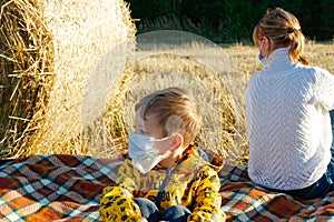 Mom and baby sit on the ground on the background of haystacks in medical masks