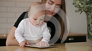 Mom and and baby sitting at the table playing tablet computer pressing your fingers on the screen and laughing