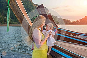 Mom and baby are standing on a pontoon pier next to wooden boats. The kid is studying mom with interest. Early morning, sunrise on