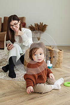 Mom and baby toddler in room. Girl is playing. Mother is using smartphone