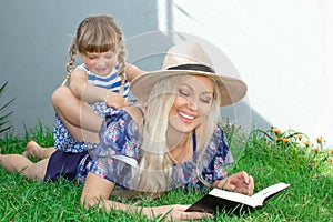 Mom blonde in a hat and her daughter are lying on the grass and reading a book, happy family.