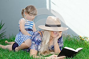 Mom blonde in a hat and her daughter are lying on the grass and reading a book, happy family.