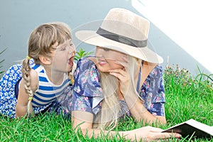 Mom blonde in a hat and her daughter are lying on the grass and reading a book, happy family. photo