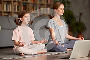 Mom with child little daughter doing yoga online, sitting in lotus pose on floor and meditating with closed eyes