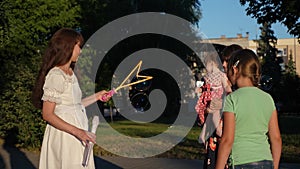 Mom and children are blowing bubbles in city park. Nanny entertains teenagers and little baby on an evening walk