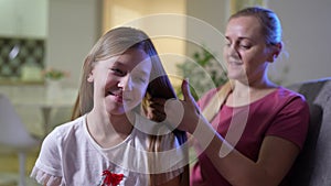 Mom is combing her daughter`s hair in the evening while sitting on the couch at home