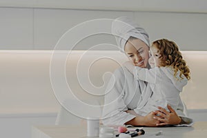 Mom and cute little daughter in white bathrobes hugging and enjoying weekend together at home