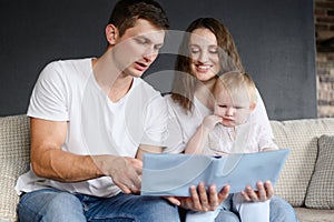 Mom, dad and child are sitting on sofa at home, reading together, looking at funny educational pictures in book. Family