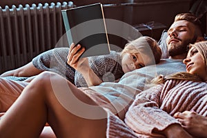 Mom, dad and daughter reading storybook together while lying on bed.