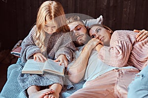 Mom, dad and daughter reading storybook together while lying on bed.