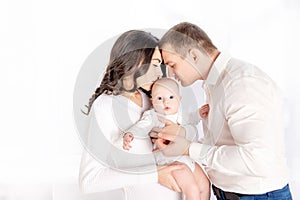 Mom and dad kiss a newborn baby holding it in their arms at home by the window, the concept of a happy loving family, lifestyle