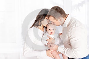 Mom and dad kiss a newborn baby holding it in their arms at home by the window, the concept of a happy loving family, lifestyle