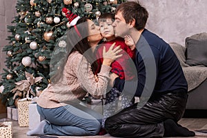 Mom and dad kiss a young son near the Christmas tree. Happy New Year`s morning with the family. Parents and child