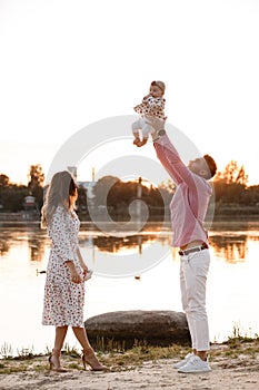 Mom, dad lifts high his adorable baby girl up mid air and looks at her smiling. Happy parents spending time playing with daughter