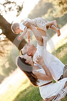 Mom, dad lifts high his baby boy up mid air and looks at her smiling. Happy parents spending time playing with son in park on