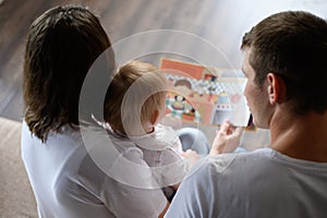 Mom and dad read an educational book to a child, sitting on the sofa, looking at pictures, rear view. Young loving