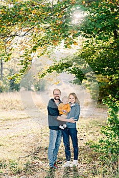 Mom and dad stand hugging with a little boy in their arms in the park