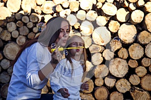 Mom and daughter blow bubbles on the background of firewood. Happy woman with child blow bubbles near stacked firewood.