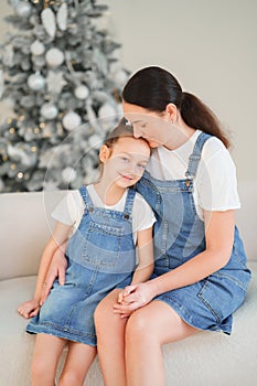 Mom and daughter in denim sundresses hugging against of a Christmas tree.