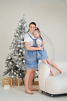 Mom and daughter in denim sundresses hugging against of a Christmas tree.