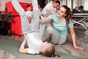 Mom and daughter do gymnastic exercises at home on the rug. Dad and son are sitting on the sofa in the background