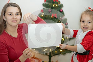 Mom and daughter dressed in red hold a white sheet of paper against the background of the Christmas tree