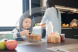 Mom and daughter eating Cereals with milk having breakfast in kitchen.