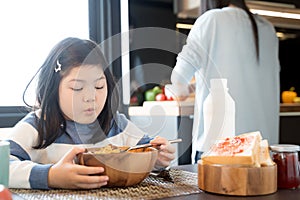 Mom and daughter eating Cereals with milk having breakfast in kitchen.