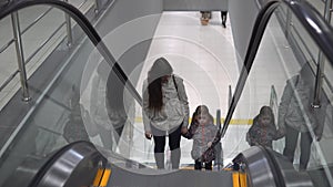 Mom and daughter are holding hands while standing on the escalator climbing up