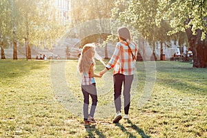 Mom and daughter holding hands walking in the park, golden hour, back view