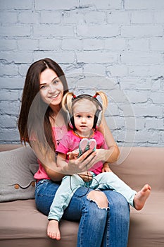 Mom and daughter listen to music in big headphones put on their head, sitting on the sofa. Holds the phone.