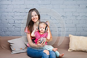 Mom and daughter listen to music in big headphones put on their head, sitting on the sofa. Holds the phone.