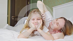 mom and daughter are lying in bed. mom adjusts her daughter's hair.