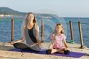 Mom and daughter are meditating on a pier near the sea, sitting on a mat in a lotus position, on a sunny summer evening