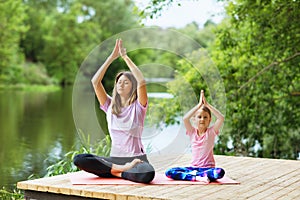 Mom and daughter perform a yoga exercise on the river bank on a warm summer day