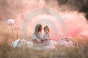 Mom and daughter in pink fairy-tale dresses are sitting in a field surrounded by Big pink decorative flowers