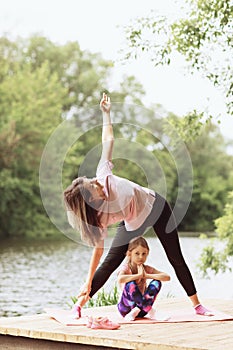 Mom and daughter practice yoga asanas on a wooden bridge near the river on a warm sunny day. Healthy lifestyle concept