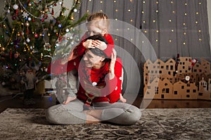 Mom and daughter in a red sweater for Christmas at the Christmas tree. A girl and her mother are hugging, kissing at the Christmas