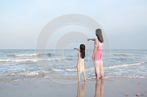 Mom and daughter relaxing on the beach on vacation. Mother pointing something with child girl looking in the sea. Back view