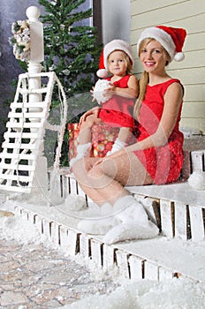 Mom and daughter in Santa`s suits are sitting under snow