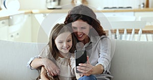 Mom and daughter sit on sofa hold smartphone having videocall